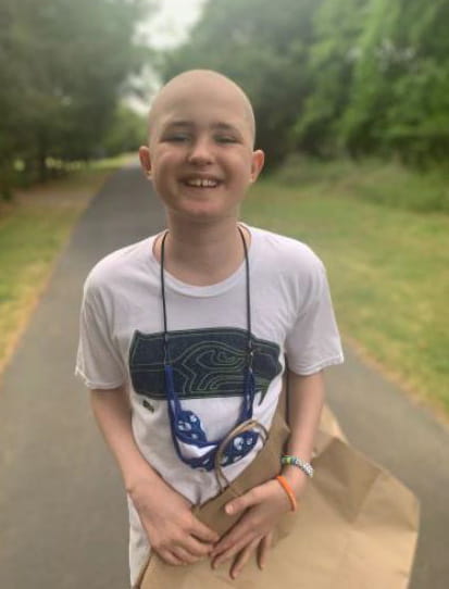 A former cancer patient beams at the camera. The teenage boy is bald and wearing a T-shirt and a face mask around his neck. He is holding a brown paper bag.