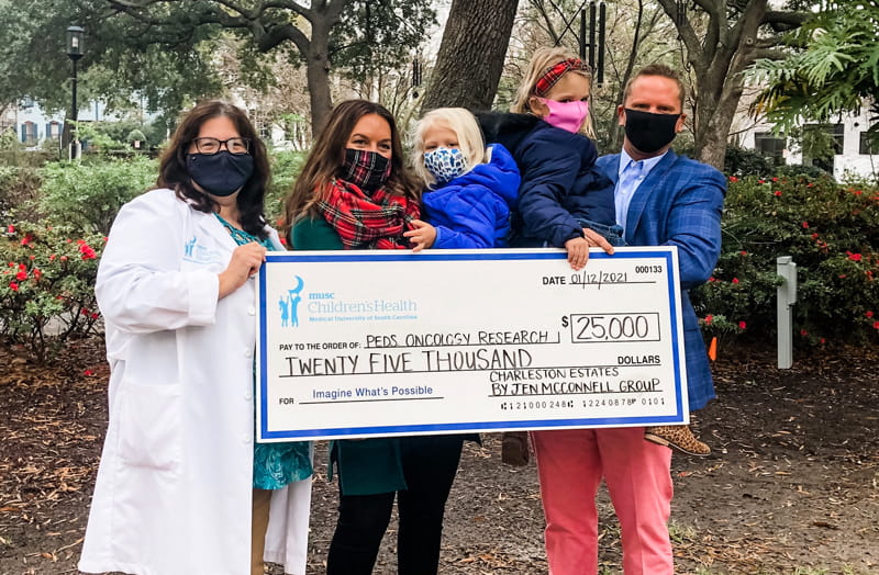 Dr. Kraveka and the McConnell family, all wearing face masks, hold a giant check for $25,000. The family has committed to raising $50,000 for Kraveka’s lab in 2021.