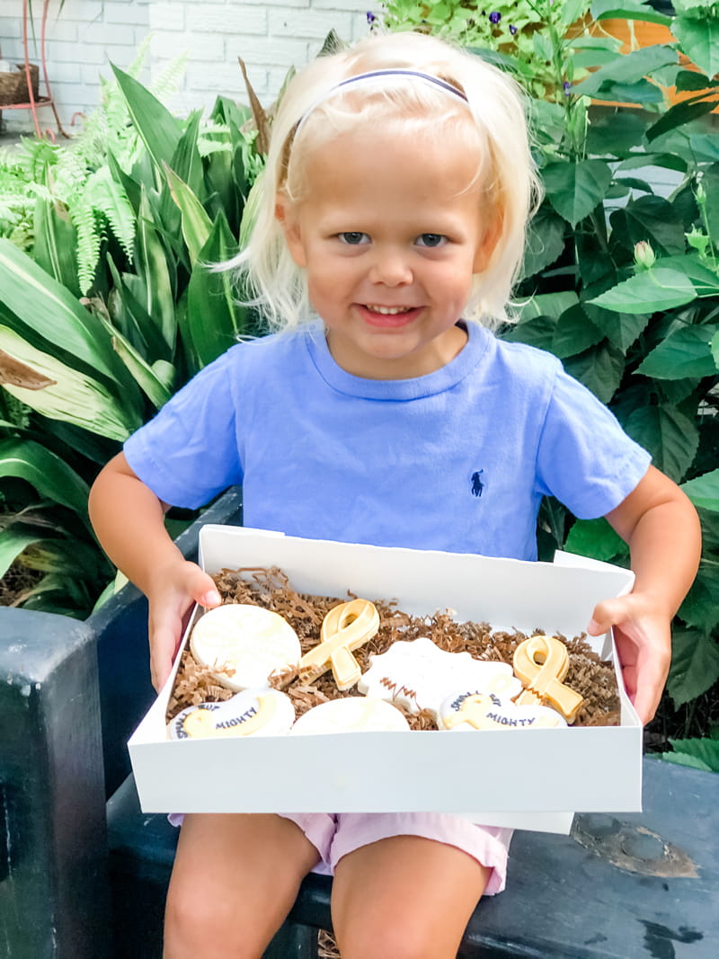 Bodhi, with shoulder length white-blond hair pulled back in a headband, holds a box of doughnuts in the shape of cancer awareness ribbons.