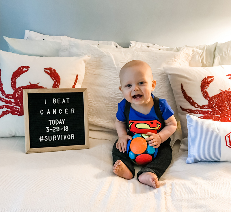 Bodhi, wearing a Superman shirt and jeans with suspenders, is propped up in front of pillows on a bed. To his left is a letterboard sign. In all capital letters, it reads, “I beat cancer today. 3-29-18. #survivor” 