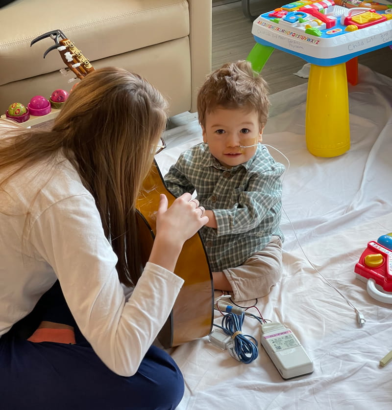 A music therapist sits kneels on a blanket on a hospital floor, holding a guitar while a 2-year-old heart touches the strings and smiles for the camera. She is wearing blue scrub pants and a long-sleeved white T-shirt. He is wearing a green plaid shirt and khakis and has a tube and wires attached to him.