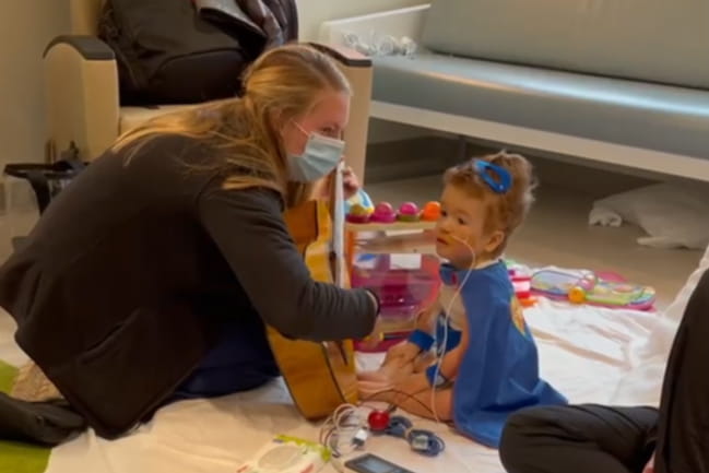 A music therapist kneels on a blanket on a hospital floor, playing guitar for a 2-year-old heart patient dressed as a super hero. He is wearing a blue cape with blue goggles pushed up on his head.
