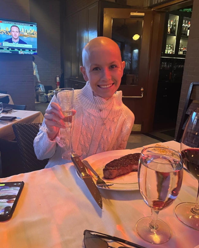 A bald teenage cancer patient smiles as she holds up her water glass. She is sitting at a table in a restaurant. There is a large steak on the plate in front of her. 