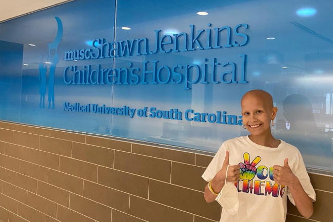 A bald teenage cancer patient poses in front of a blue sign that reads, MUSC Shawn Jenkins Children’s Hospital, Medical University of South Carolina. She has a big smile and holds two thumbs up. She’s wearing a white T-shirt, with a tie-dyed peace symbol and lettering that reads, “Peace out chemo.”