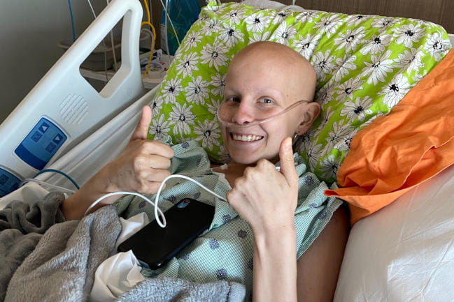 A bald teenage cancer patient poses in her hospital bed with two thumbs up. Her head rests on a pillow with a bright green flower-patterned pillowcase, and she has a feeding tube in. 