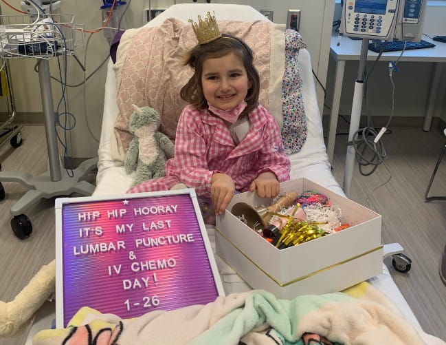 A young girl in pink gingham pajamas sits up in a hospital bed. She has a gold crown on and a message board that reads: Hip Hip Hooray. It’s my last lumbar puncture and IV chemo day. 1-26.