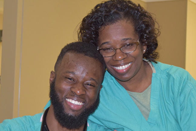 Mother and son, both nurses wearing scrubs, pose for a photo in a hospital. Both have big smiles. 