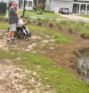 A preteen boy in a wheelchair fishes in a neighborhood pond. He is accompanied by an adult. 