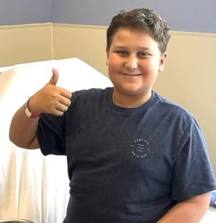 A preteen boy with brown hair sits on a hospital bed and gives a thumbs up.