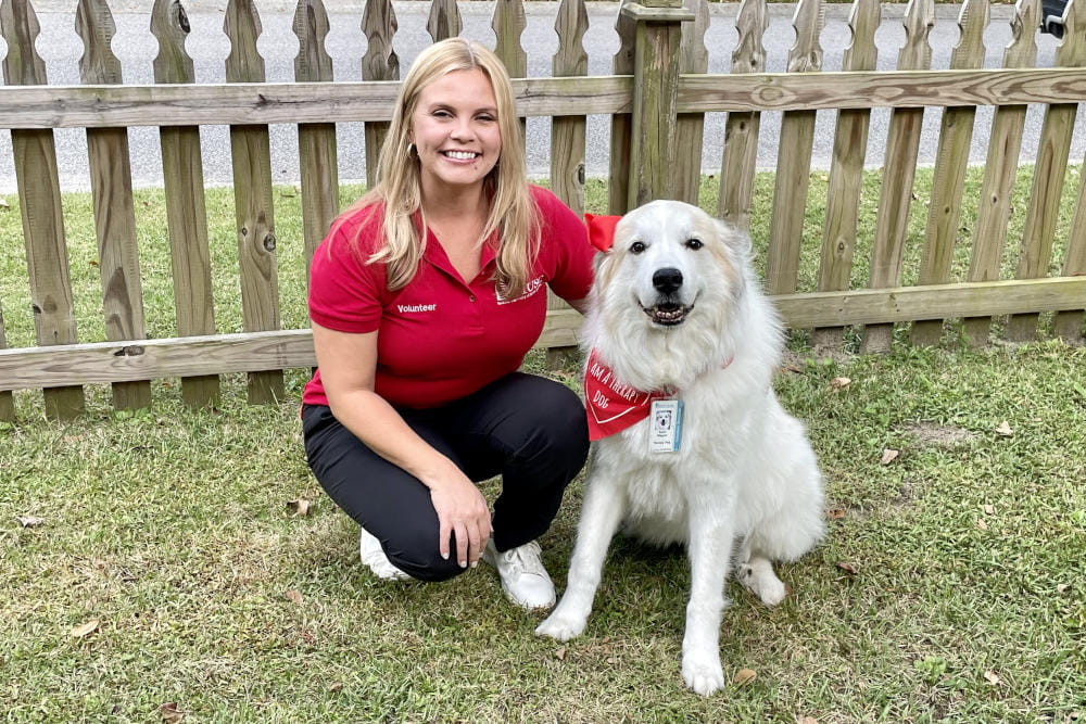 An animal therapy volunteer in a red shirt and a canine volunteer in a red bandana kneel in a yard in front of a fence.