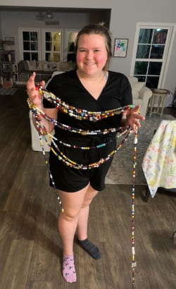 A person stands indoors, smiling, holding multiple long beaded necklaces. The background features a living room with wooden flooring, furniture, and a large window