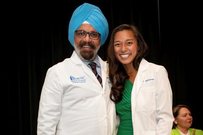 A female student in a green dress and white coat, poses for a photo with the college’s dean, also dressed in a white coat.