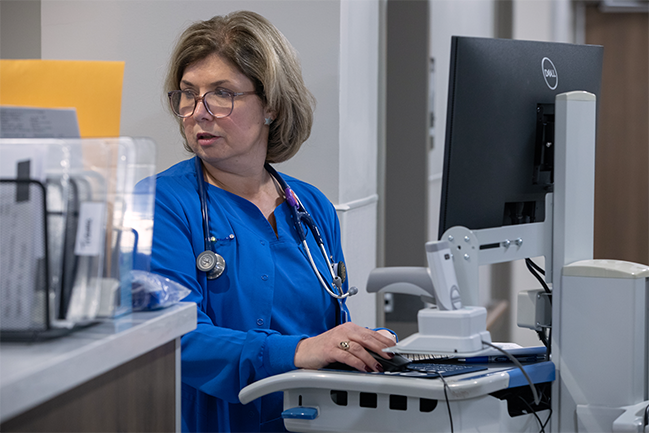 A nurse in blue scrubs, with a stethoscope around her neck, types on a computer in a hospital setting. She appears focused and attentive. 