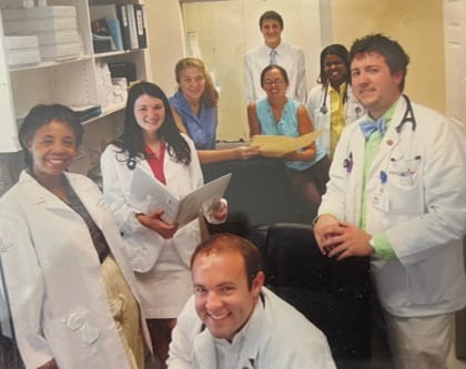 A team of health care professionals, including doctors and nurses, posing together with smiles in a clinical environment.