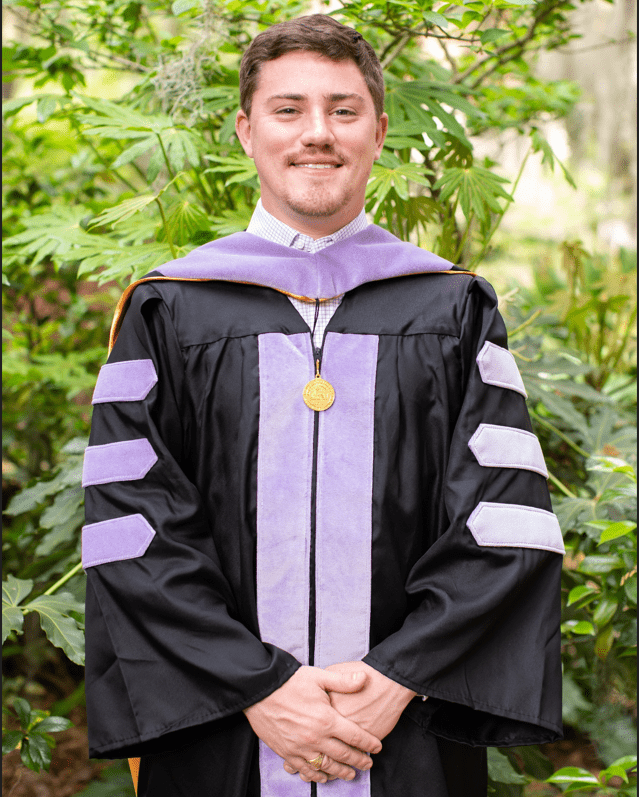 A man in a graduation robe and purple tie, standing confidently as he marks a significant milestone in his education. 