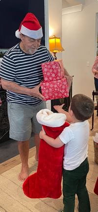 An older man in a Santa hat gives gifts to a child holding a large red stocking.