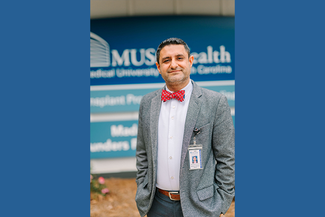 A man in a gray blazer and red bow tie stands confidently with a slight smile. He has a badge with a photo ID clipped to his blazer. In the background is a blurred blue sign reading "MUSC Health." 