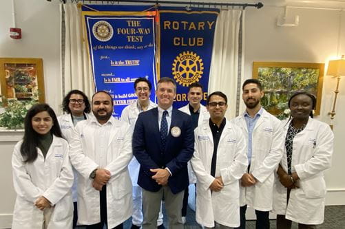  A group of doctors smiling for a photo in front of a rotary club banner, showcasing their professional camaraderie.