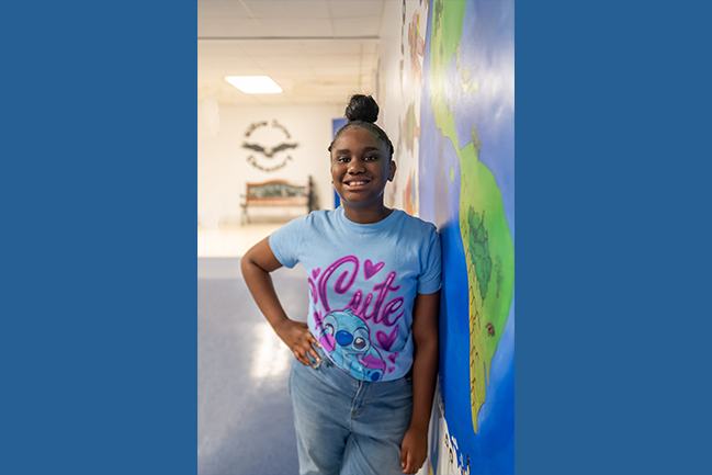 The image features a person standing indoors, leaning slightly against a brightly painted wall. The person is smiling and wearing a light blue t-shirt with a graphic design featuring a cartoon character and the word "Cute" in pink. They are also wearing blue jeans. 