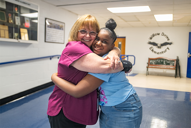 The image shows a cheerful scene of a middle-aged woman and a young girl embracing in a hallway. The woman is wearing glasses and a purple shirt, while the girl is dressed in a light blue T-shirt and jeans. They are both smiling at the camera.   