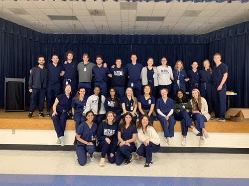 A group of people in medical scrubs and casual clothing from the MUSC College of Medicine pose proudly on a stage in a school gymnasium.