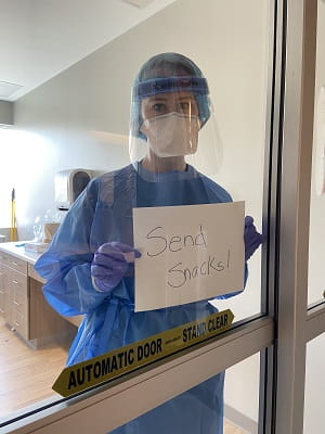 A nurse in personal protective equipment stands in front of an interior window holding a sign that reads, ‘Bring snacks.