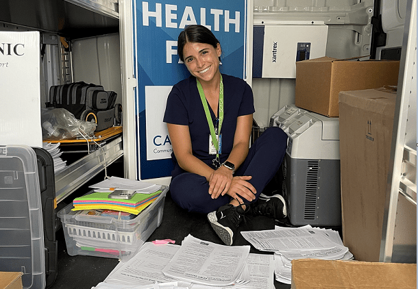  Person sitting on the floor of a van surrounded by documents and boxes, wearing a lanyard and navy scrubs. A large "Health" sign is visible in the background. 