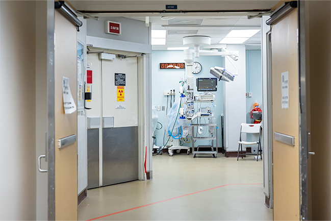 The image shows the interior of a hospital or medical facility corridor leading into a room with medical equipment.  