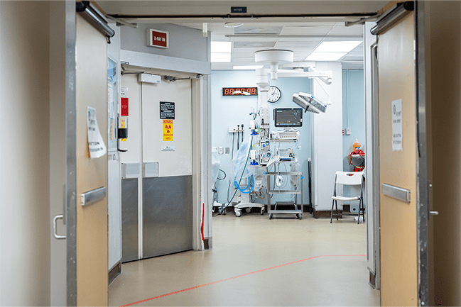 The image shows the interior of a hospital or medical facility corridor leading into a room with medical equipment.  