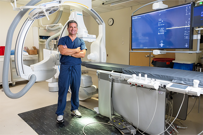 Medical professional in blue scrubs in a room with advanced imaging equipment and a large C-arm machine. 