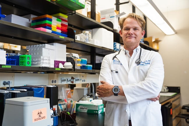 A man in a lab coat stands confidently in front of shelves filled with various scientific equipment and supplies.