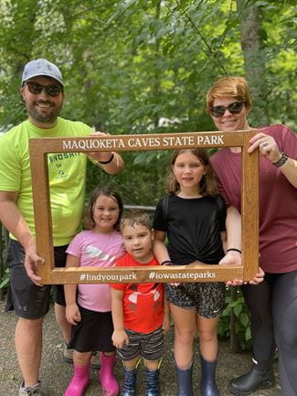 A family poses with a wooden frame labeled "Maquoketa Caves State Park" in a forest setting. Transcribed text: MAQUOKETA CAVES STATE PARK #findyourpark #iowastateparks 