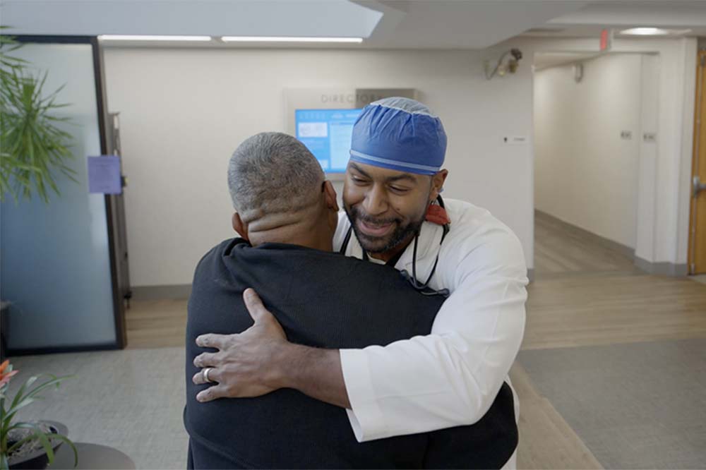 A man in a blue cap and a white doctor's coat hugs another man whose back is to the camera.